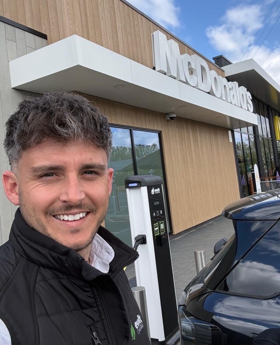 A man facing the camera stands beside an EV which is plugged into an Evolt Charging EV charger at a McDonald's parking lot.